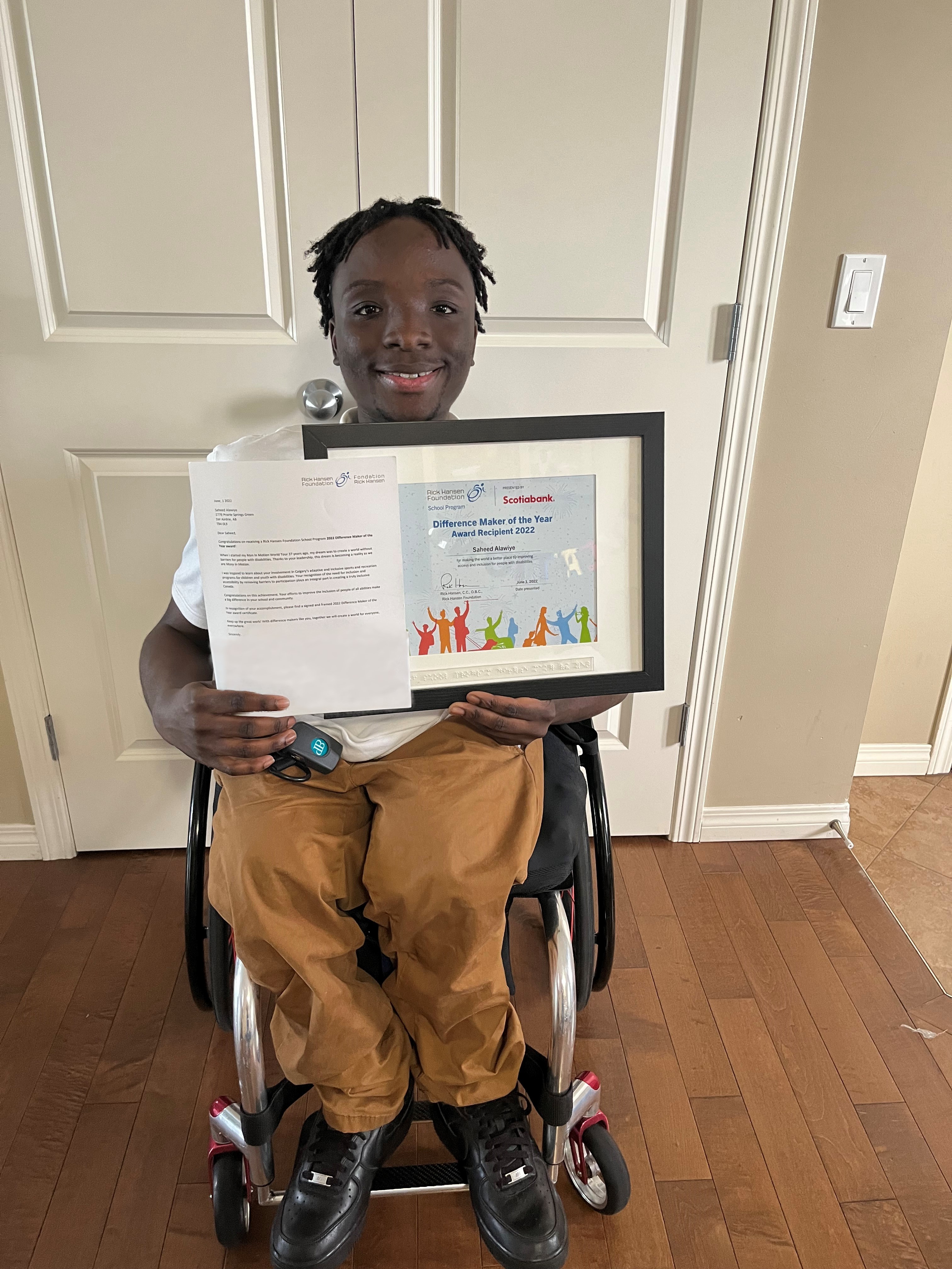 Saheed who is using his wheelchair holding his Difference Maker of the Year Award certificate. He is smiling and wearing tan-coloured pants.